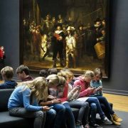 Group of teenagers looking their smart phones seated beside "The Nightwatch" classical painting in a museum.