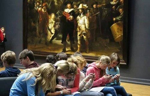 Group of teenagers looking their smart phones seated beside "The Nightwatch" classical painting in a museum.
