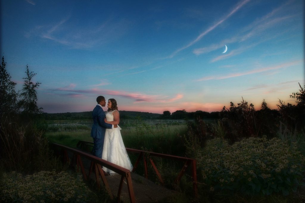 Bride and groom holding each other around the waist, standing on a footbridge against a scenic sunset.