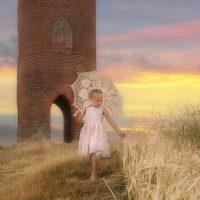 Young girl in summery dress with a parasol, walking towards camera touching grass with her hand. Brick build folly in background.