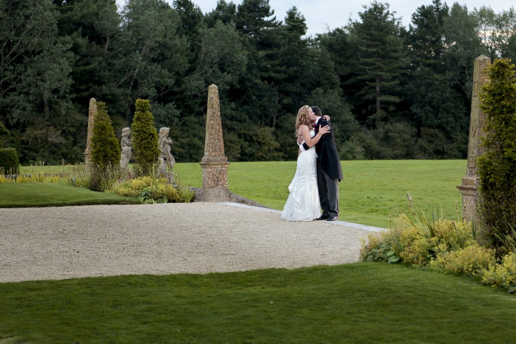 Bride and groom hugging each other standing on the path at edge of a formal garden with stone obelisk. 