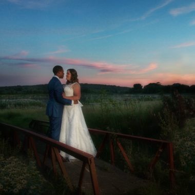 LR Wedding_5214b_1x1 Newly married couple standing on a footbridge together against a sunset.