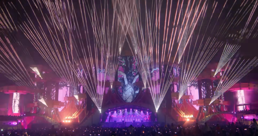 Audience view of pop concert stage, showing lots of lasers fanning out from the facade around the stage which is also lit with projected images.