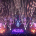 Audience view of pop concert stage, showing lots of lasers fanning out from the facade around the stage which is also lit with projected images.