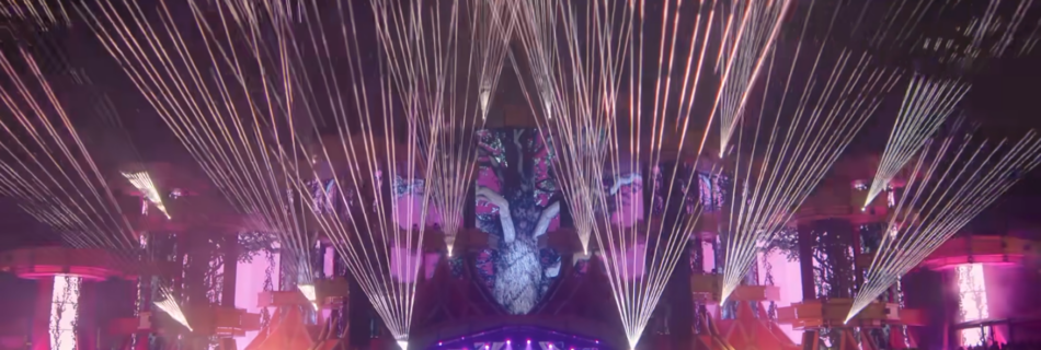 Audience view of pop concert stage, showing lots of lasers fanning out from the facade around the stage which is also lit with projected images.
