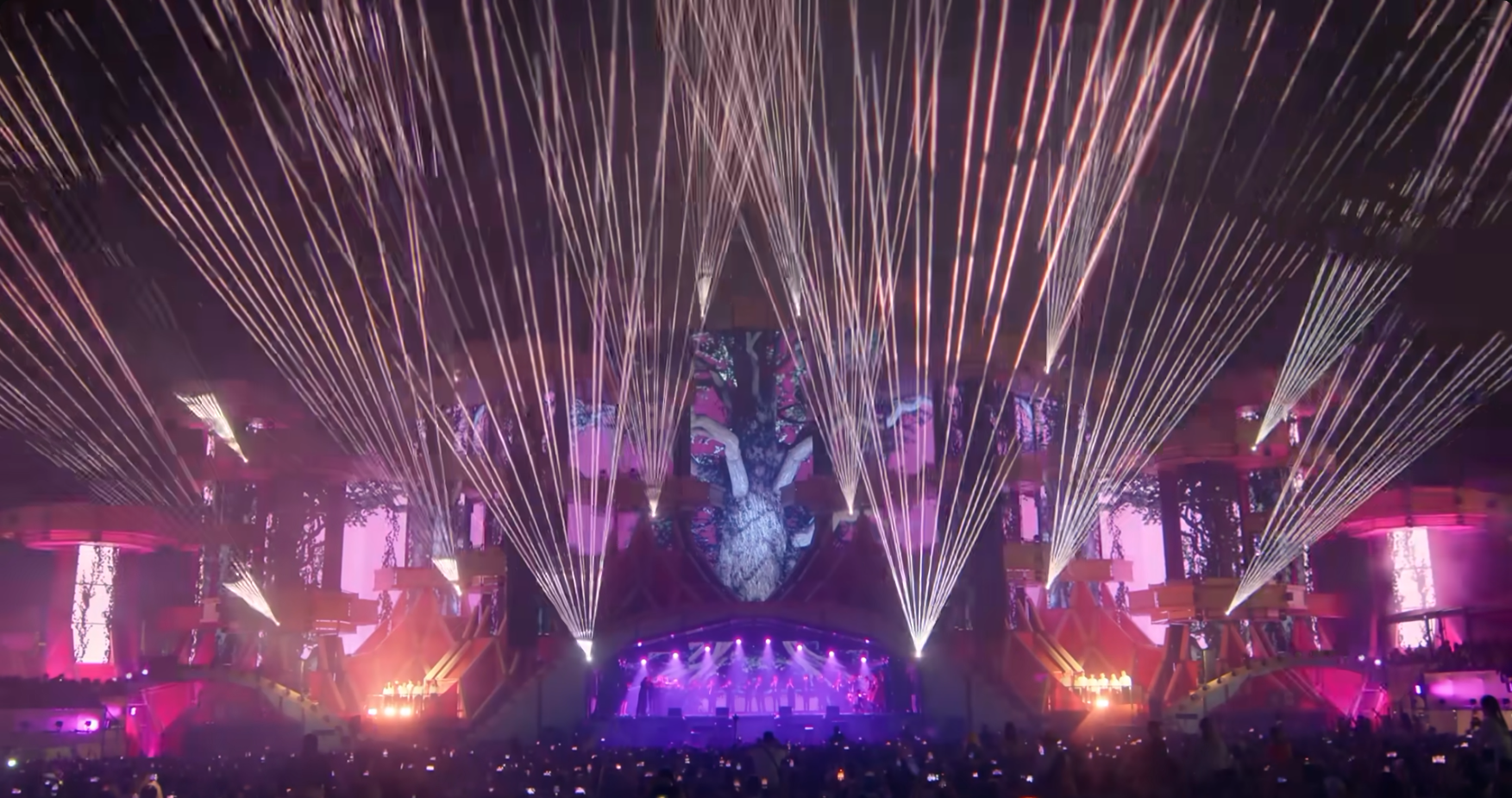 Audience view of pop concert stage, showing lots of lasers fanning out from the facade around the stage which is also lit with projected images.