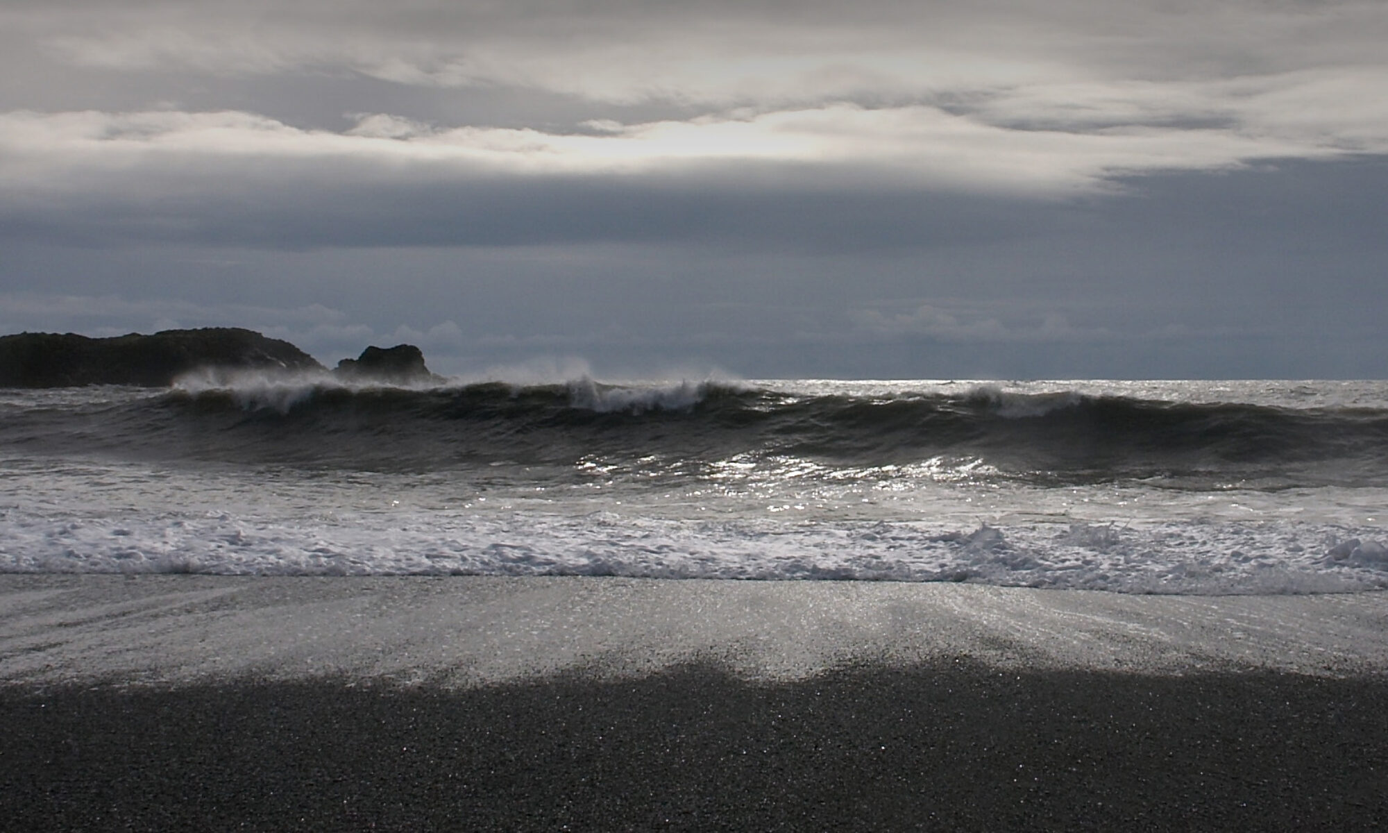 Photo taken in the early evening with a low sun behind clouds, light reflecting from rolling waves washing on a black sand beach.