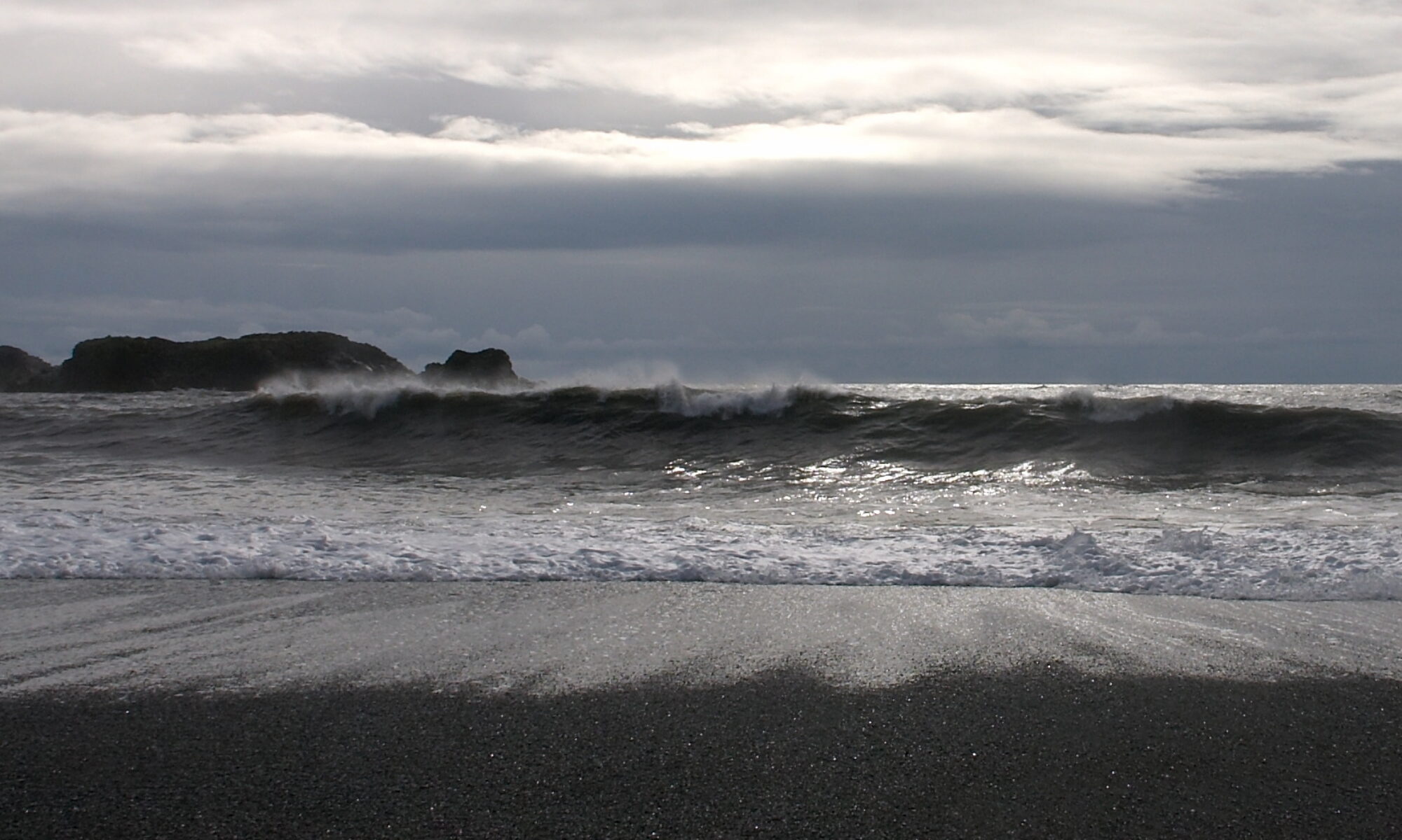 Photo taken in the early evening with a low sun behind clouds, light reflecting from rolling waves washing on a black sand beach.